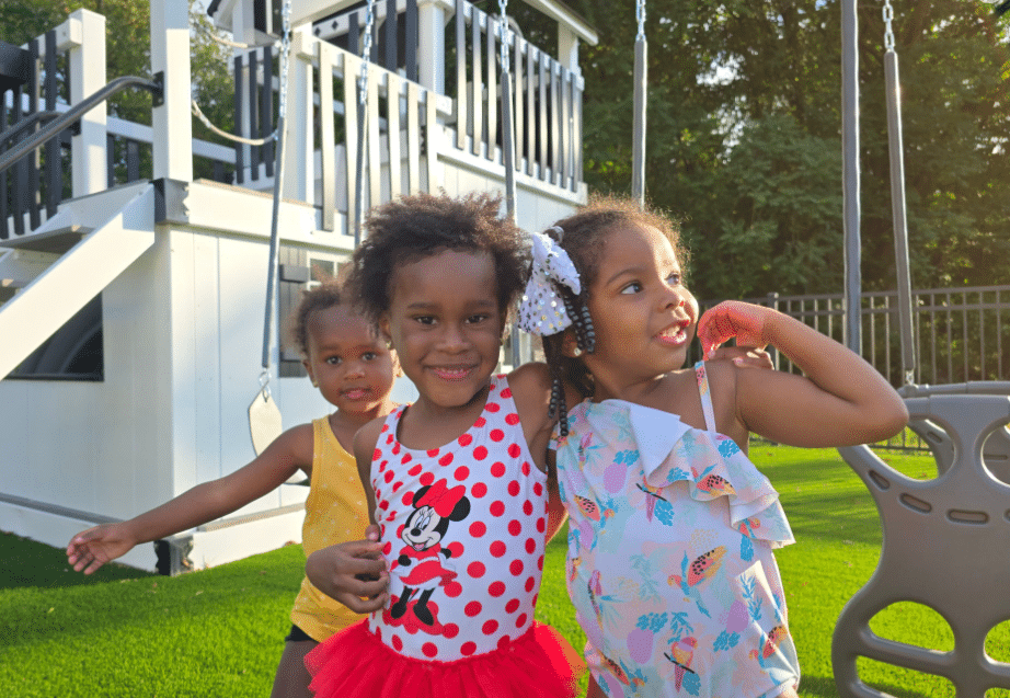 Three young children smiling and playing on a backyard with vibrant artificial turf, surrounded by a swing set and a white deck staircase in the background."