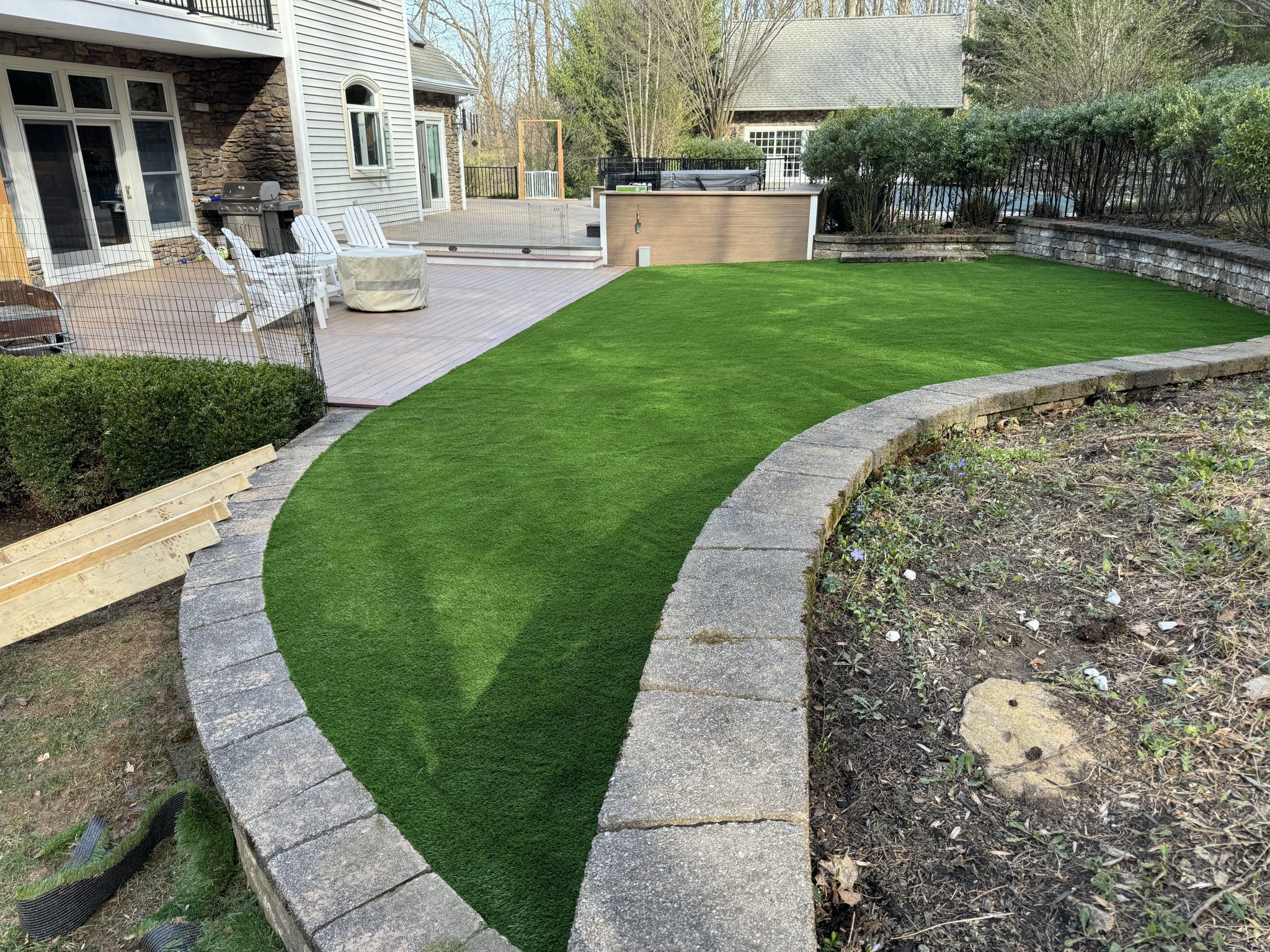 Freshly installed artificial turf in a curved backyard surrounded by a stone retaining wall, adjacent to a patio area with outdoor seating and a hot tub.