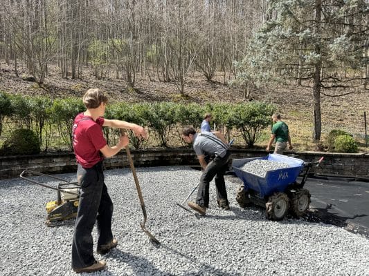 Team of workers spreading and leveling crushed stone during artificial turf installation in a backyard, surrounded by trees, shrubs, and landscaping tools