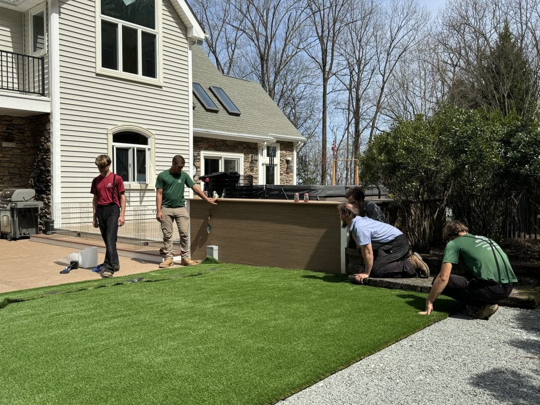 Workers installing artificial turf in a backyard, carefully aligning the edges near a patio and hot tub, with a modern house and surrounding greenery in the background.