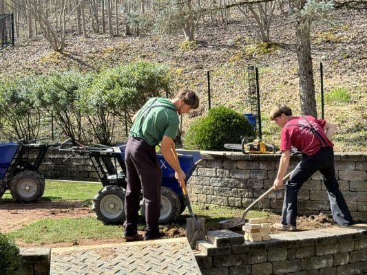 Two workers preparing a backyard for artificial turf installation by excavating the ground near a stone retaining wall, surrounded by landscaping tools and equipment